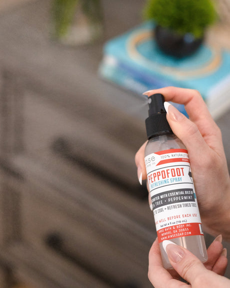 A hand holding Peppofoot Refreshing Spray beside a bedside table with a plant and stacked books — the last step before bed for tired feet. Rinse Bath & Body Co.
