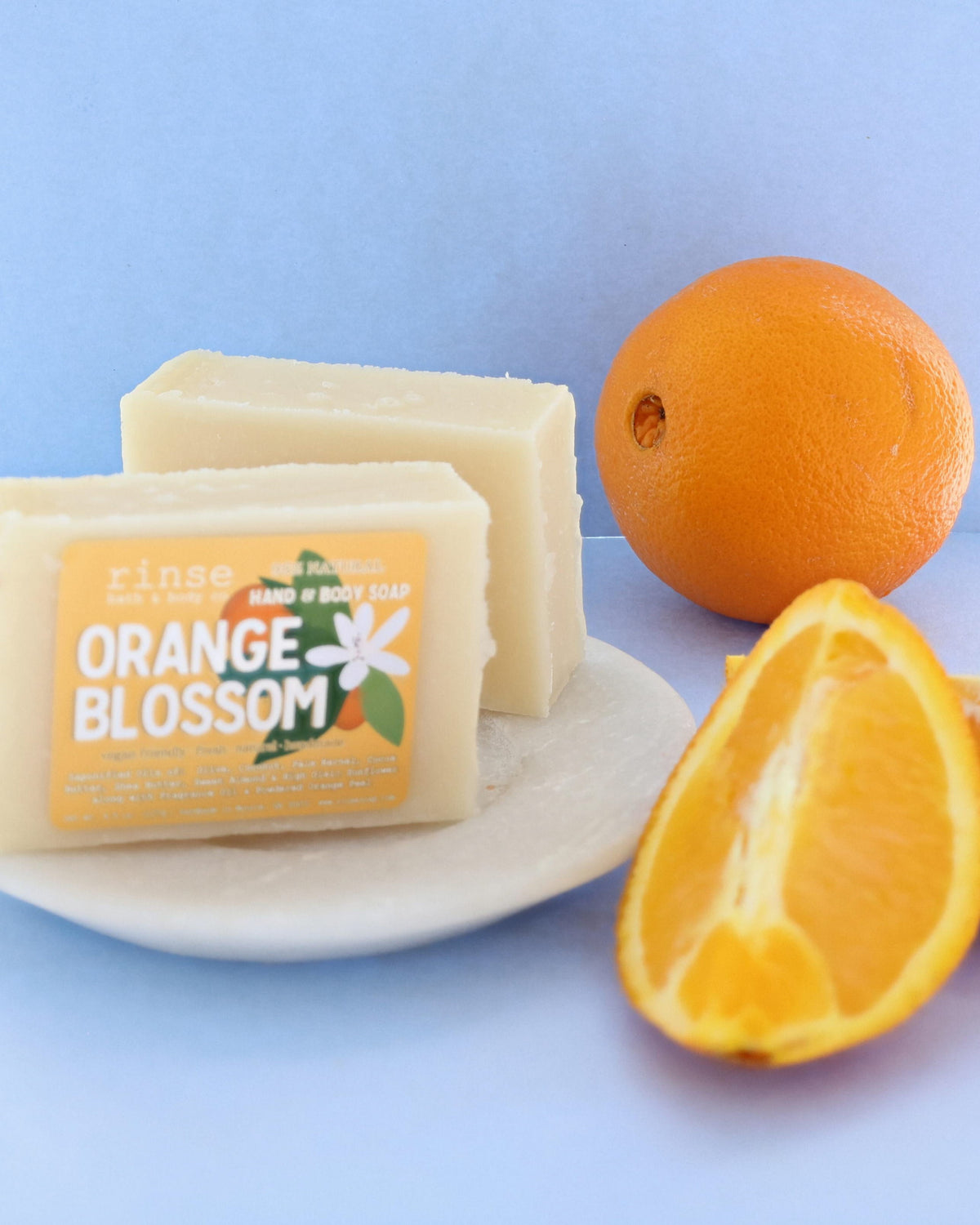 Two Orange Blossom bars on a ceramic dish with whole orange and juicy wedge — pastel blue backdrop