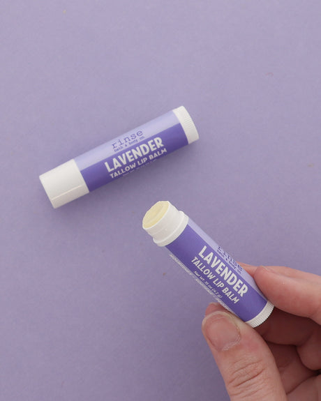 Hand holding an uncapped Lavender Tallow Lip Balm beside a capped tube on a plain lilac backdrop
