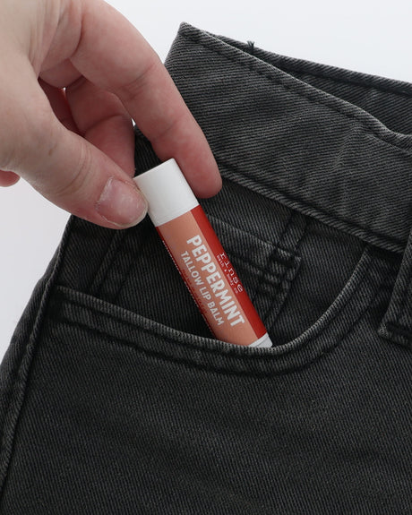 Peppermint Tallow Lip Balm half-tucked into the front pocket of charcoal jeans, held between two fingers