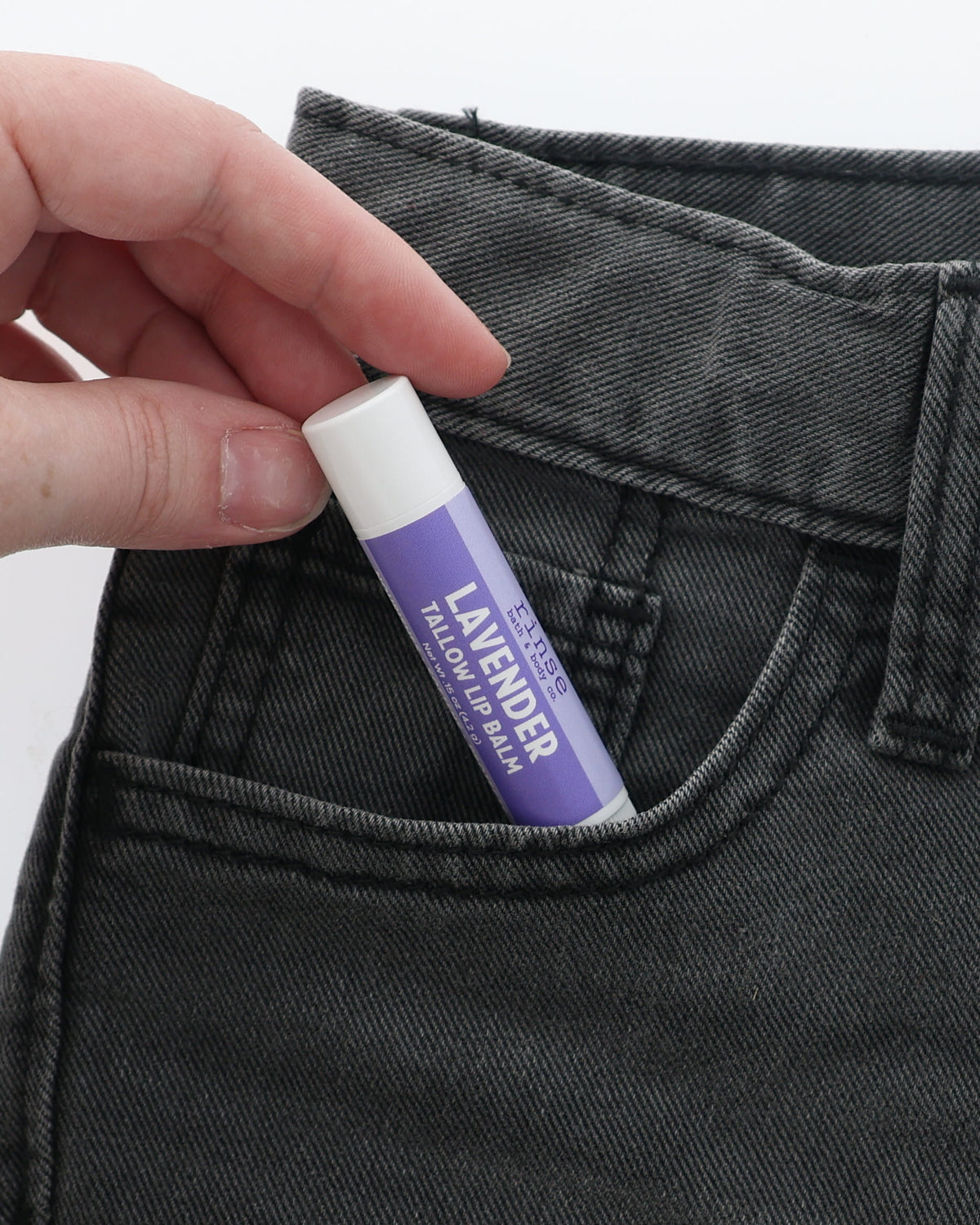 Lavender Tallow Lip Balm half-tucked into the front pocket of charcoal jeans, held between two fingers