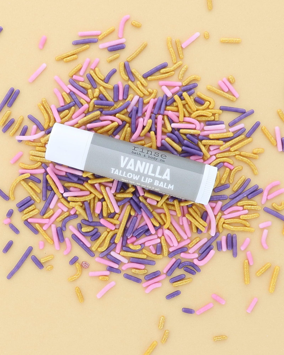 Vanilla Tallow Lip Balm tube with grey label lying on pink, purple and gold sprinkles on a butter-yellow backdrop