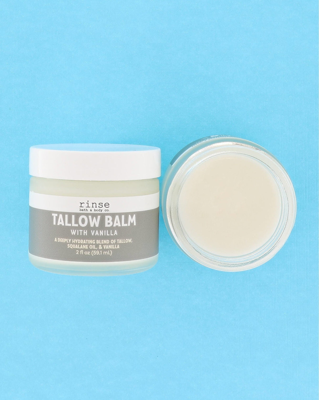 A jar of Rinse Tallow Balm with vanilla next to its open lid, showing the creamy off-white balm inside, set against a solid light blue background.