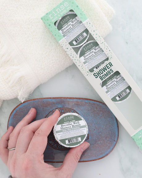 Hand sets a foil-wrapped Breathe Well shower bomb on a blue-grey stoneware dish beside the 4-pack box, a cool blend of peppermint, eucalyptus, thyme, sage and camphor. Shown for display; pack contains four (4) shower bombs.