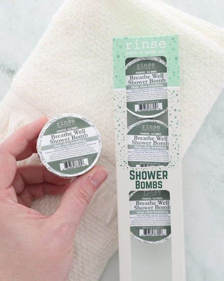 Hand lifts a Breathe Well shower bomb above a soft white towel next to the 4-pack box, ready to release peppermint, eucalyptus, thyme, sage and camphor in the steam. Shown for display; pack contains four (4) shower bombs.