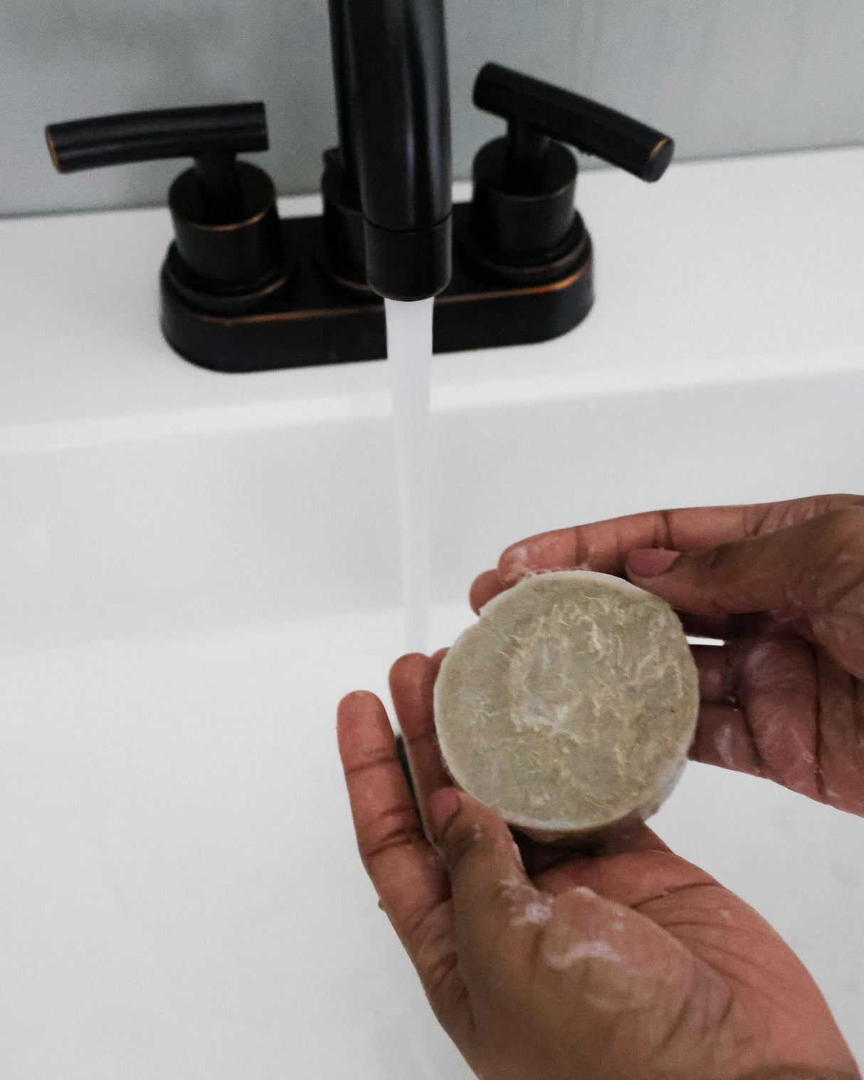 Hands lathering the round Dead Sea Mud Loofah bar under a black faucet, grey suds visible — mineral mud with a scrubby loofah back. Rinse Bath & Body Co.