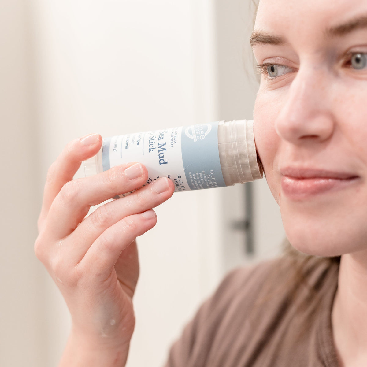 Woman smiling while gliding Dead Sea Mud Soap Stick across her cheek in bright bathroom light