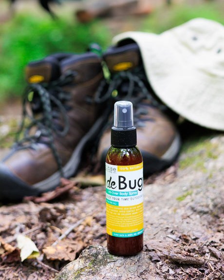 DeBug spray standing beside worn hiking boots and a canvas sun hat on a forest trail