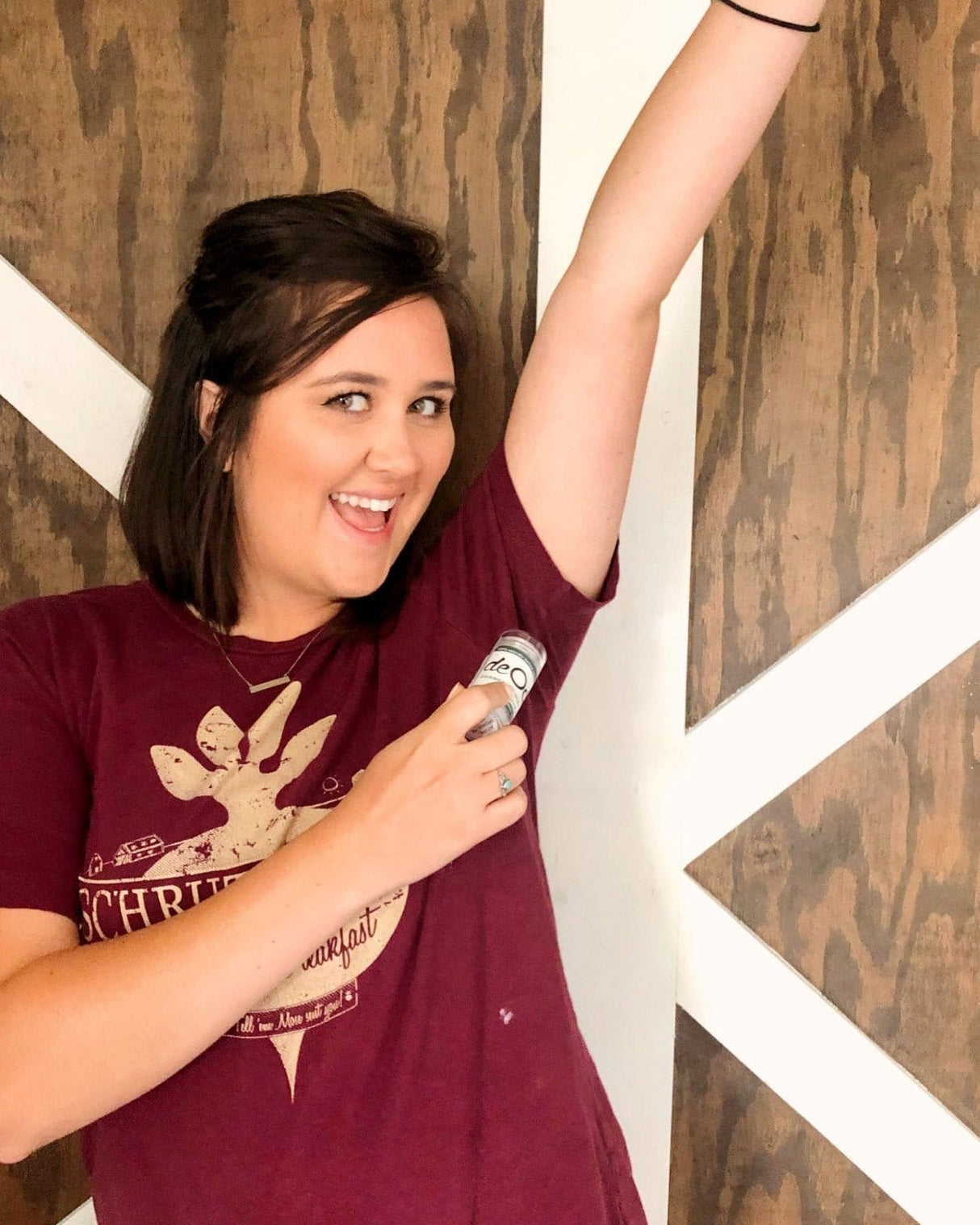 A woman in a maroon tee, arm raised, swiping a Rosemary Mint DeOdor Stick under her arm against a white and wood wall