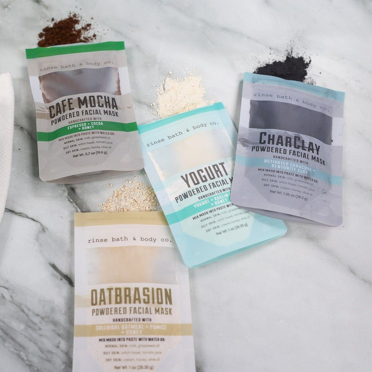 Four powdered facial mask pouches with matching powder piles on marble — Cafe Mocha, Charclay, Yogurt, and Oatbrasion textures in soft spa light