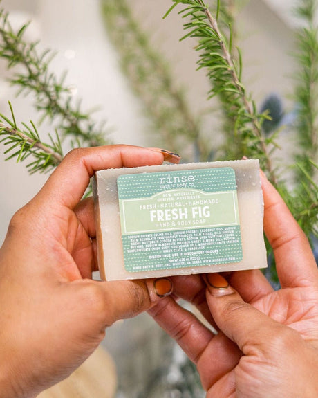 Hands holding Fresh Fig soap with sage-green clay top — soft herbal sprigs, in-use sensory moment