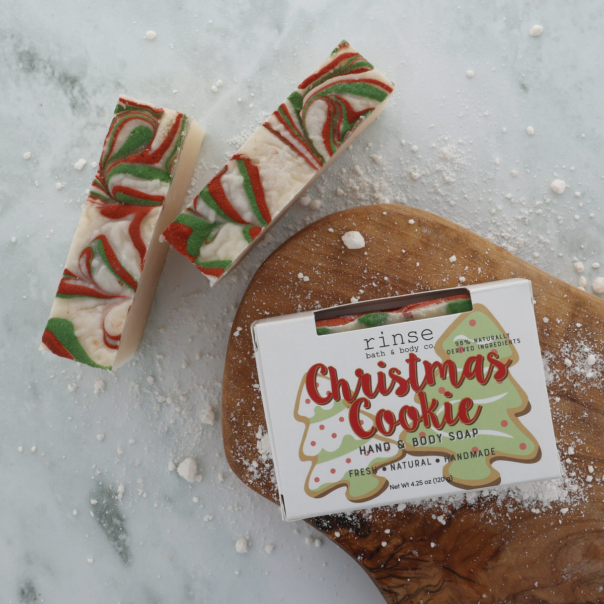 Christmas Cookie soap slices on white marble beside a frosted gingerbread cookie and a dusting of sugar — warm vanilla, butter and cinnamon, the bar that smells like the oven door opening. Rinse Bath & Body Co.
