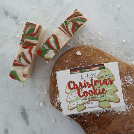 Christmas Cookie soap slices on white marble beside a frosted gingerbread cookie and a dusting of sugar — warm vanilla, butter and cinnamon, the bar that smells like the oven door opening. Rinse Bath & Body Co.