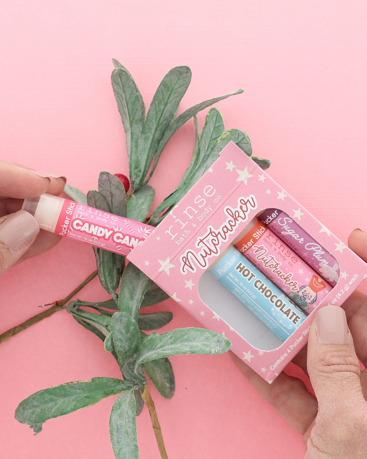 Hand holding the pink Nutcracker lip balm pack and a Candy Cane Pucker Stick with a frosted greenery sprig on pink