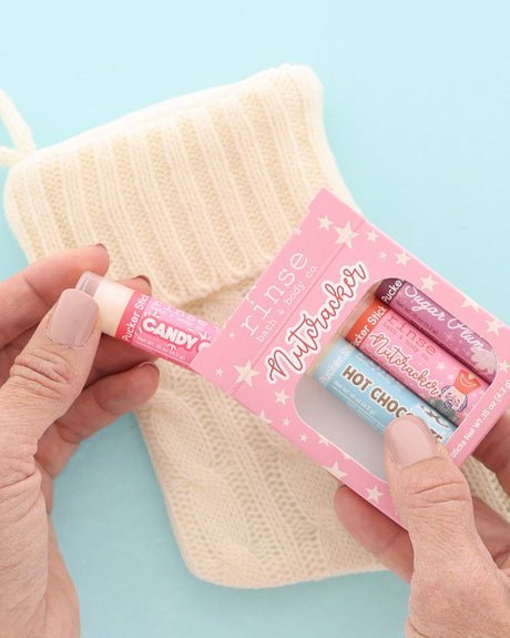 Hand pulling a Candy Cane Pucker Stick from the pink Nutcracker lip balm pack beside a cream knit stocking