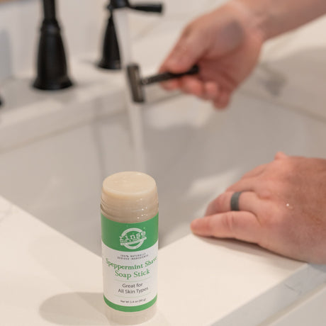 Open Speppermint Shave Soap Stick on marble with water-dotted label — razor rinsing under a black faucet in bathroom light