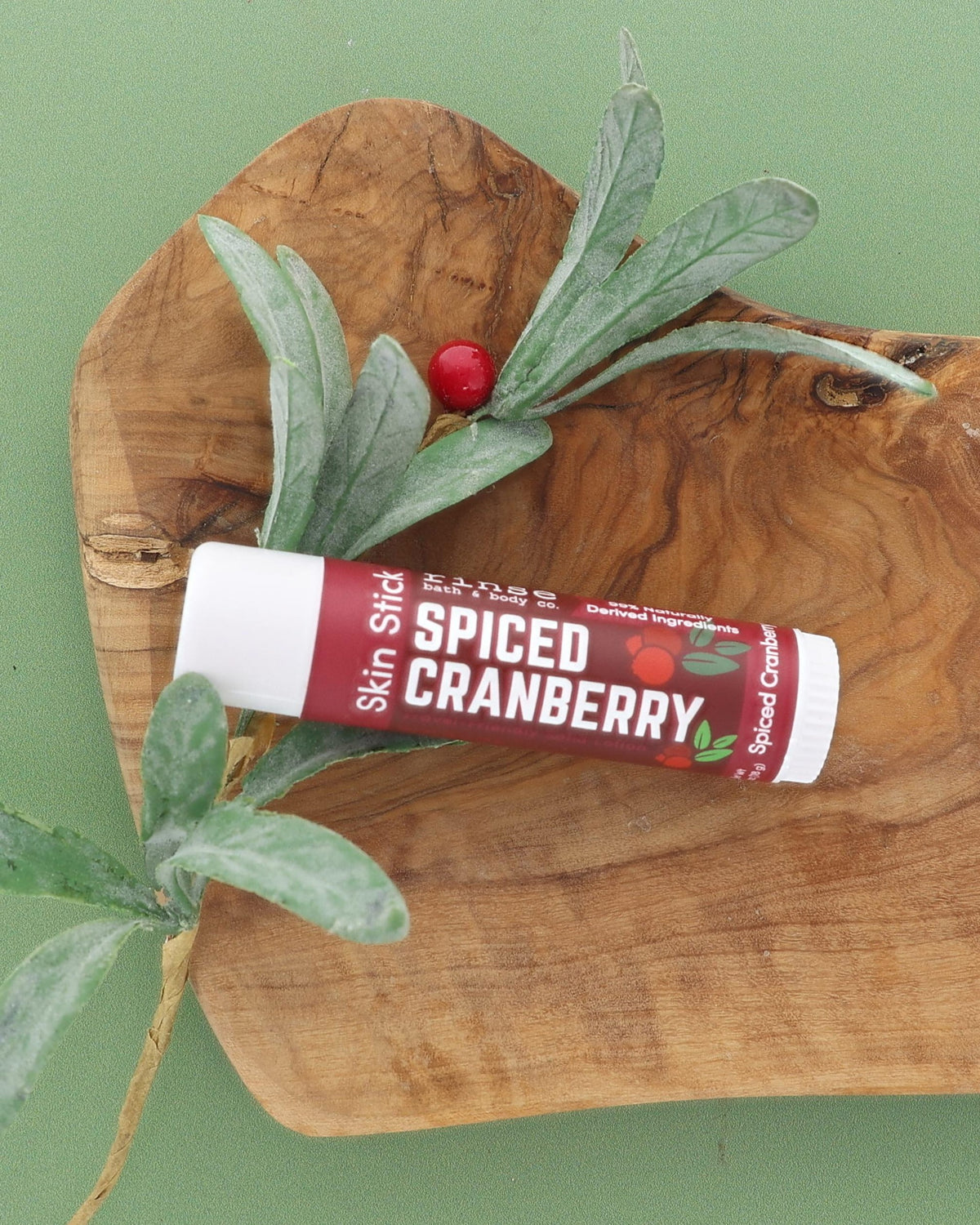 Spiced Cranberry Skin Stick on an olive wood board with a frosted greenery sprig and red berry on sage green