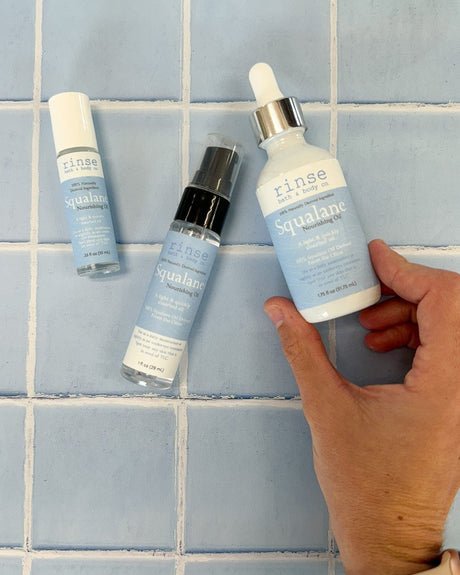 Hand holding large Squalane dropper beside roller and spray bottles — cool blue tile grid in bright vanity light