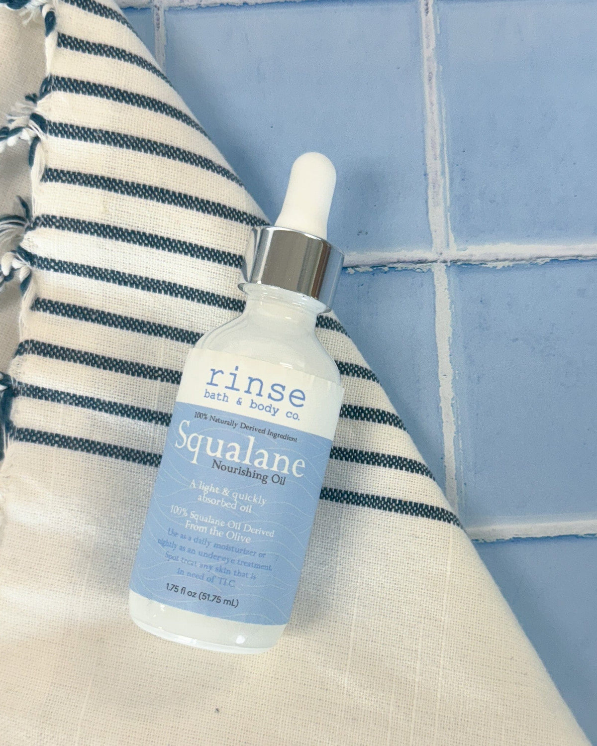 The 2 oz Squalane Face Oil dropper bottle standing on a tiled ledge beside a striped towel. Rinse Bath & Body Co.