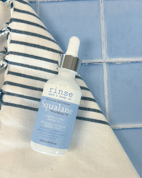 The 2 oz Squalane Face Oil dropper bottle standing on a tiled ledge beside a striped towel. Rinse Bath & Body Co.