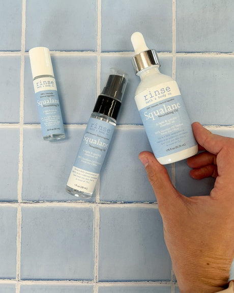 A hand lifting the Squalane Face Oil dropper from a group of Rinse oil bottles arranged on blue tile. Rinse Bath & Body Co.