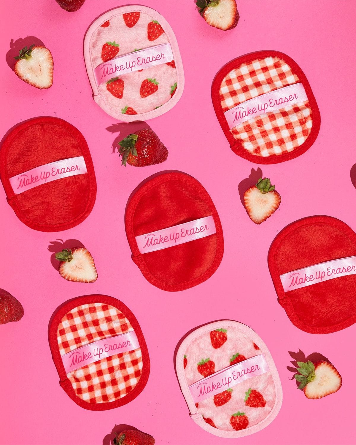 Seven mini Strawberry Fields MakeUp Eraser cloths scattered on pink with fresh strawberries — red, gingham, and strawberry-print pads