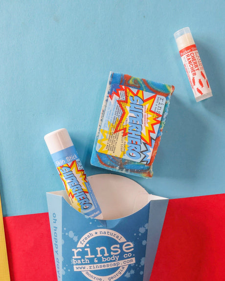 The Superhero kids gift set styled on blue, red and yellow colour-blocked paper — rainbow-swirl bar soap, Superhero solid lotion skin stick and a Cherry Rancher lip balm beside the blue carton. Rinse Bath & Body Co.