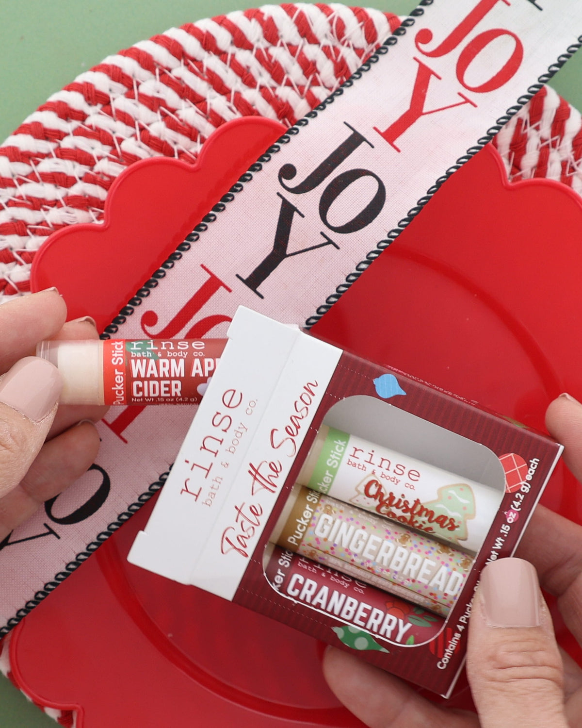 Hand holding the Taste the Season lip balm pack over a red woven plate with a JOY ribbon