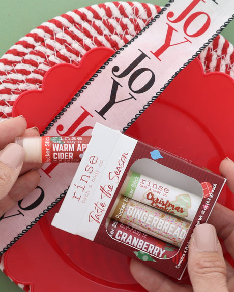 Hand holding the Taste the Season lip balm pack over a red woven plate with a JOY ribbon