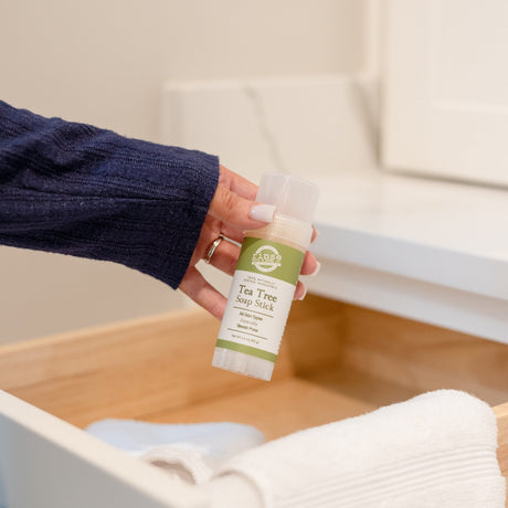 Hand lifting a Tea Tree Soap Stick from an open wooden drawer of white and blue towels — green label in bright light
