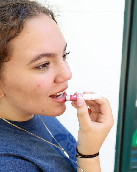 A woman in scrubs applying the Daring tinted lip balm — the deepest of the three tints. Rinse Bath & Body Co.