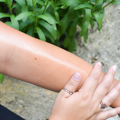 Forearm gleaming with Vanilla Honeycomb oil under leafy green blur — silver rings rest on sunlit outdoor skin