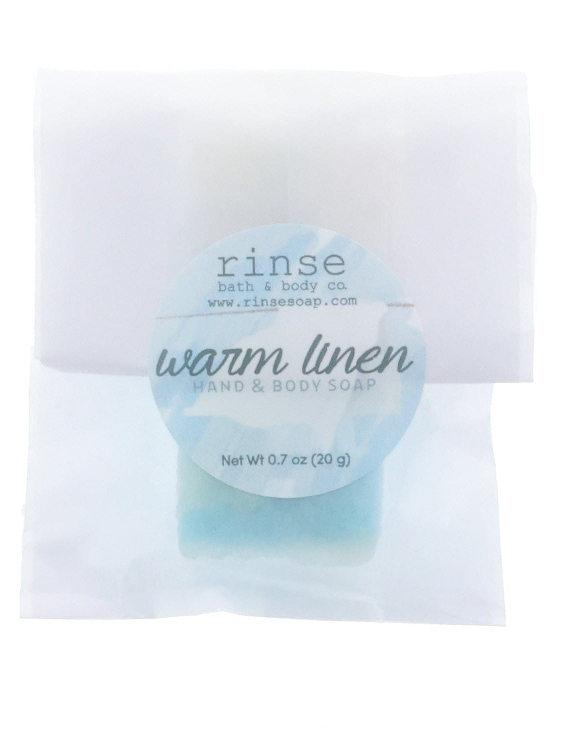 Warm Linen soap slice rests in crinkled translucent paper under a soft blue circular label on bright white.