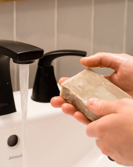 Hands working the Beard Bar into a lather at the bathroom sink — a cold-process face and beard soap with Dead Sea mud and bentonite clay. Rinse Bath & Body Co.