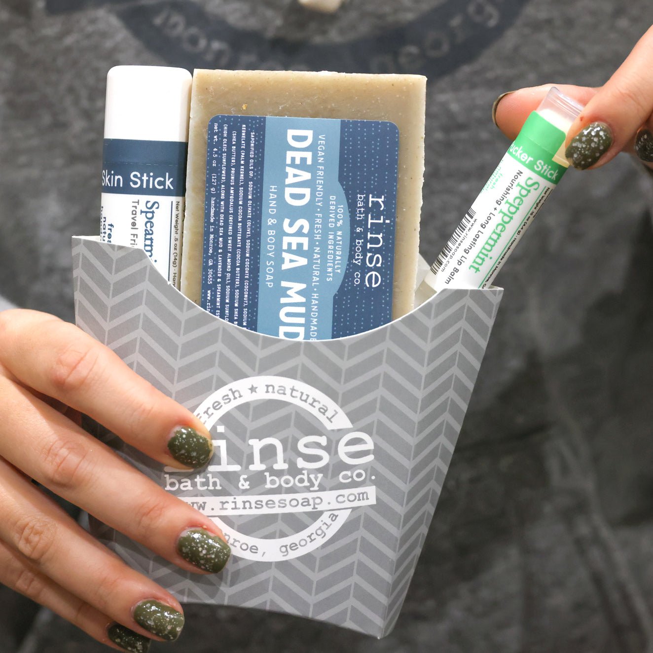 A hand holding the Dead Sea Mud fry-box gift set against concrete — Dead Sea Mud bar soap, Spearmint Lavender solid lotion stick and a Speppermint lip balm in the grey chevron carton. Rinse Bath & Body Co.