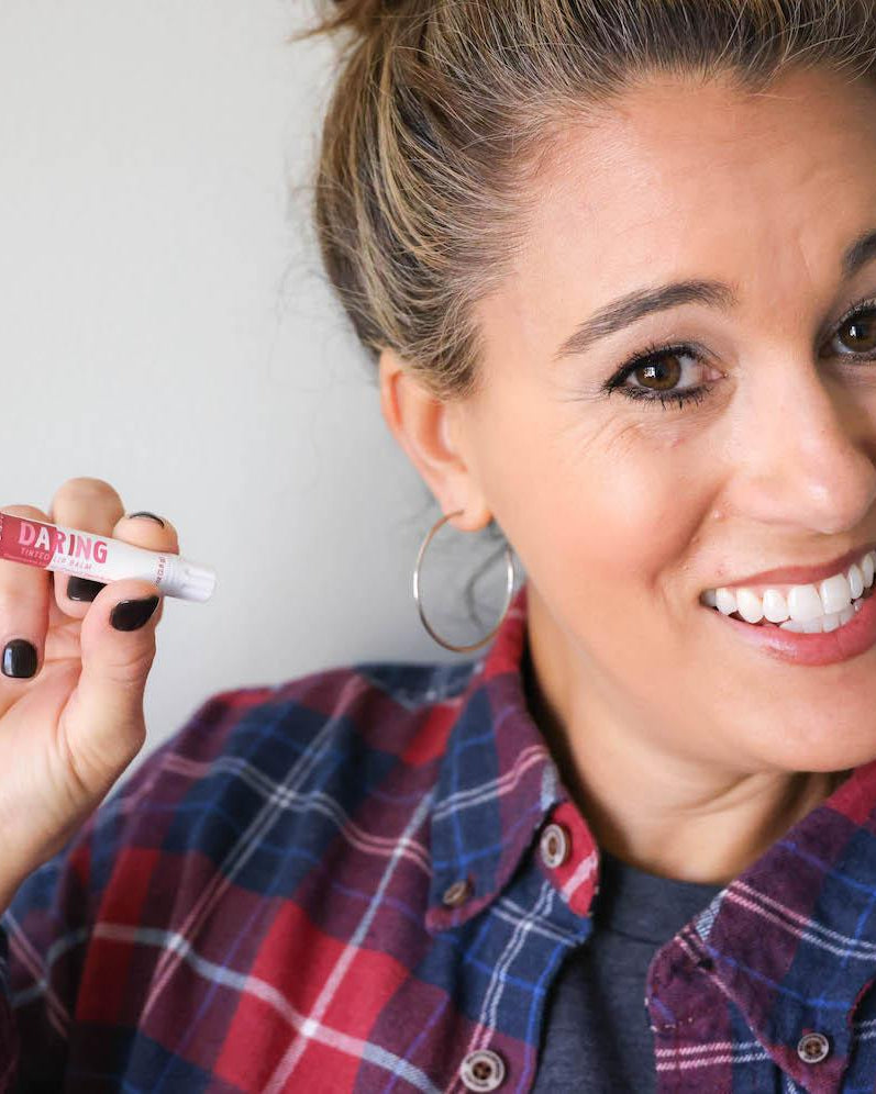 A woman in a plaid shirt holding the Daring tinted lip balm, wearing its sheer berry colour. Rinse Bath & Body Co.