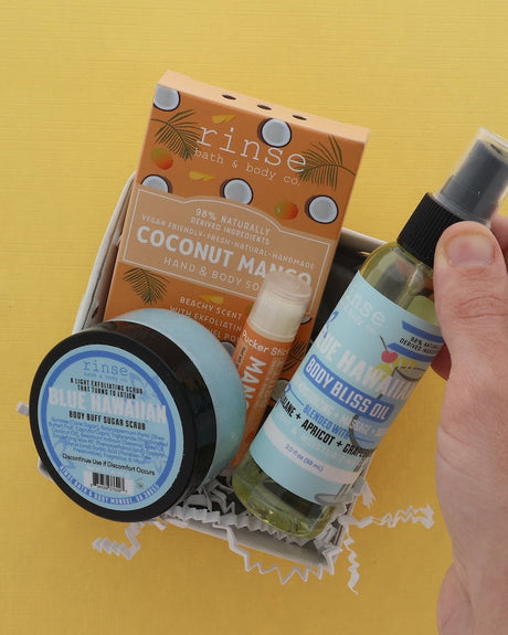 A hand lifts a white gift box on a sunny yellow background holding the four-piece Tropical Scrub & Shine set — Coconut Mango soap, Blue Hawaiian Body Buff sugar scrub, a Pucker Stick lip balm and Blue Hawaiian Body Bliss Oil. Rinse Bath & Body Co.