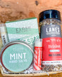 Wooden basket of Mint Hand Salve, Peppermint and Vanilla Puckers, soap, and Brisket rub on wood before brick — rustic Dad gift in daylight