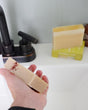 Hand rinsing Chardonnay soap under a matte black faucet — botanical flecks on cream bar in bright bathroom light