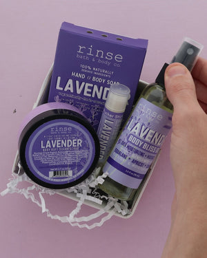 A hand holds a lavender bath and body gift set from Rinse Bath & Body Co. The set includes lavender hand & body soap, a lavender body bliss oil, a lavender body butter jar, and a lavender pucker stick  all in purple packaging inside a white gift box with white paper shred, against a solid lavender background.