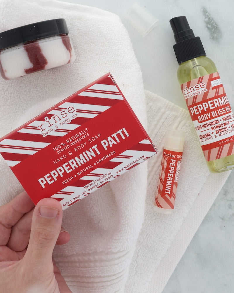 A hand holds a red and white striped box of Rinse the Scum Peppermint Patti hand and body soap next to a bottle of Peppermint Body Blossom spray and a lip balm, all arranged on a white towel over a marble surface.