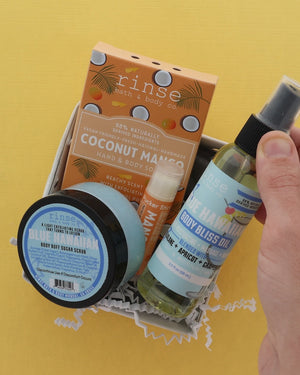 A hand holding a gift set of bath and body products from Rinse and The Handmade, including Coconut Mango hand & body soap, Blue Raspberry body butter, and Blue Bliss body oil, arranged in a white box against a yellow background.