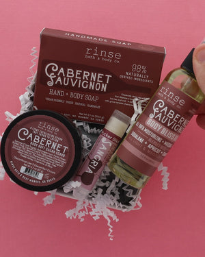A hand holds a bottle of Rinse Hand & Body Soap next to a jar and a box of Cabernet Sauvignon hand and body soap, presented in white crinkle paper on a solid pink background.