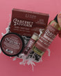 A hand holds a bottle of Rinse Hand & Body Soap next to a jar and a box of Cabernet Sauvignon hand and body soap, presented in white crinkle paper on a solid pink background.