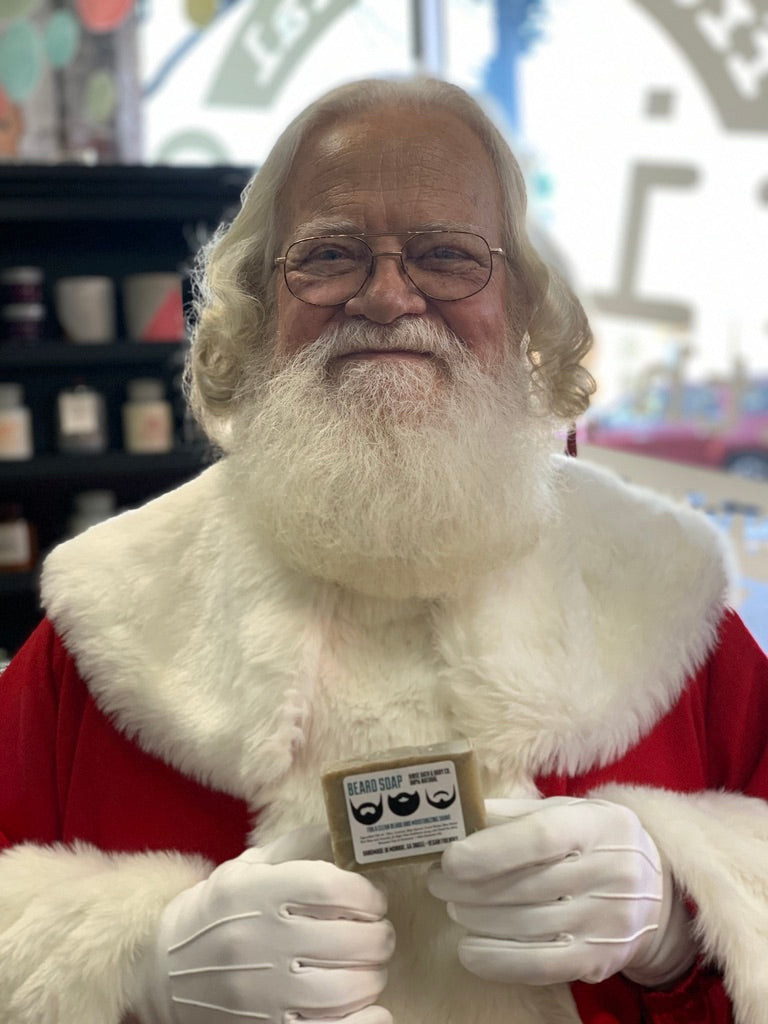 Smiling Santa in fur-trimmed red holds wrapped Beard Soap — soft window light in a shop aisle