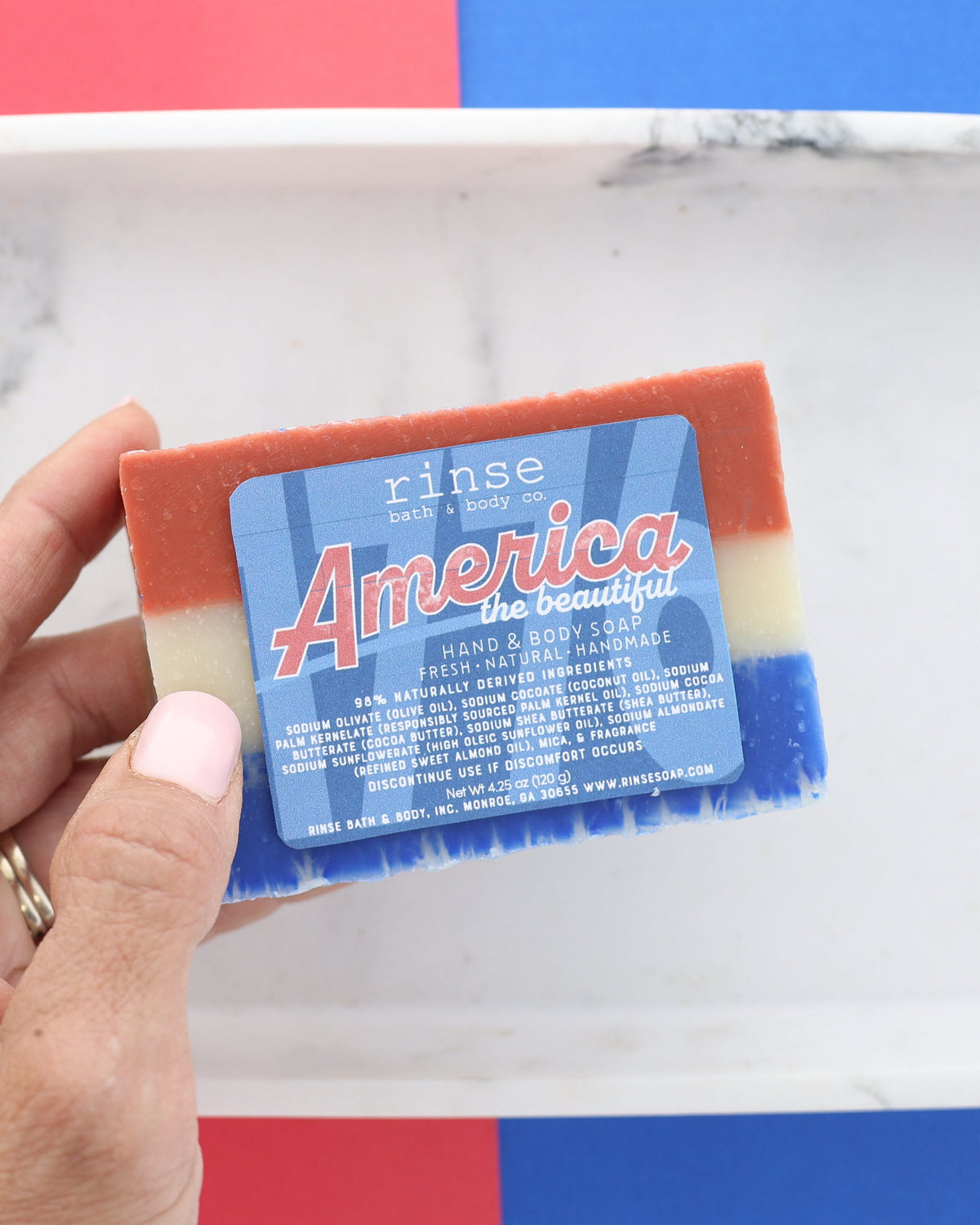 Hand holding America the Beautiful soap over a white marble tray — tri-color red, cream, and blue layers in soft daylight