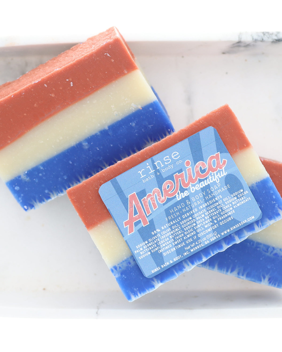 Two America the Beautiful soap bars resting on pale marble — layered red, cream, and cobalt catching clean daylight