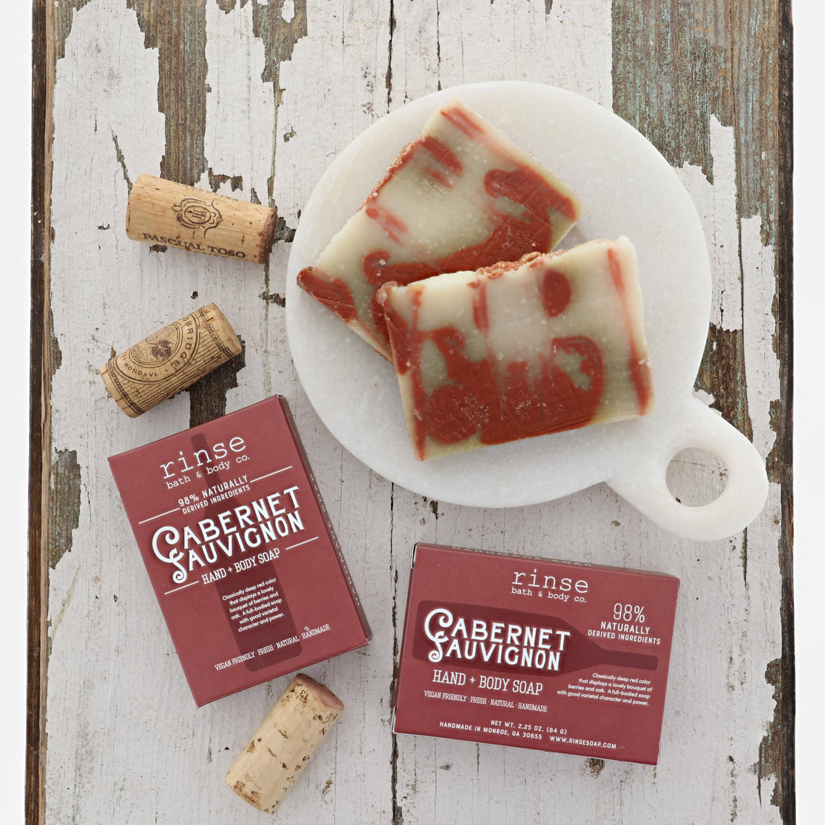 Two Cabernet Sauvignon bars on a white marble board beside corks and burgundy boxes — cream-and-wine swirls on weathered wood