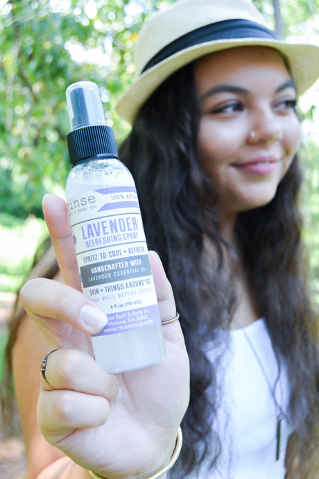 A woman in a beige hat holds up a bottle of lavender refreshing spray against a blurred outdoor green background.