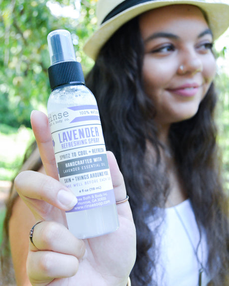 A woman in a beige hat holds up a bottle of lavender refreshing spray against a blurred outdoor green background.